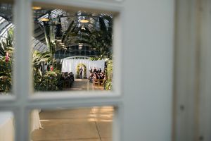A wedding ceremony viewed through a window in a lush greenhouse venue with tropical plants.