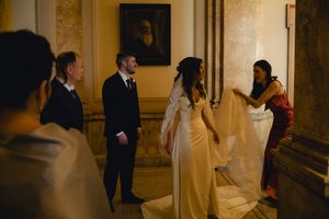 Bride in elegant white gown having veil adjusted by bridesmaid in golden-lit classical interior.