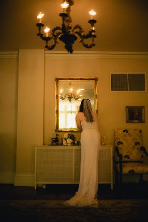 Bride in white gown and veil standing before ornate gold mirror in warmly lit elegant room.