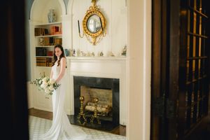 Elegant bride in white gown holding bouquet posing by fireplace in a classic interior.