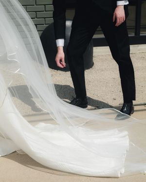 Groom in black tuxedo standing beside flowing white bridal veil train on outdoor pavement.