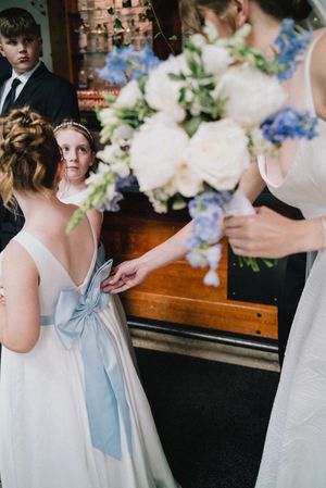 Young flower girl in white dress with blue bow holding bride's hand at church wedding ceremony.