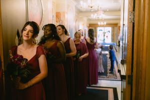 Bridesmaids in burgundy and purple gowns line a elegant hallway, with the first bridesmaid holding a floral bouquet.