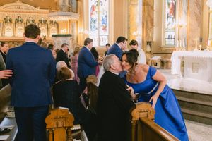 Guests greet each other warmly in a ornate Catholic church during a wedding ceremony with stained glass windows.