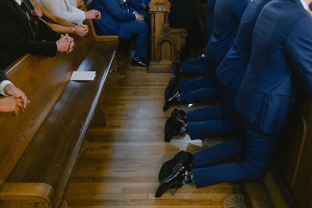 Wedding party in navy blue suits seated in wooden church pews during Catholic ceremony.