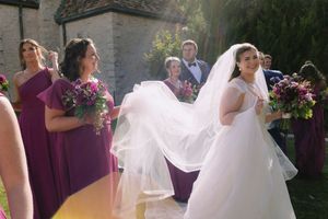 Bridesmaids in purple dresses help arrange the bride's flowing white veil outdoors at a wedding.