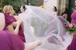Bridesmaids in magenta dresses hold and arrange the bride's billowing white veil before the ceremony.