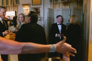 Elegantly dressed wedding guests in black tie attire mingle and converse in a warmly lit reception room.