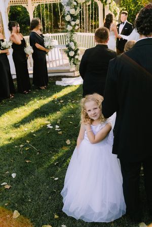 Adorable curly-haired flower girl in white tulle dress smiles during an outdoor Wisconsin garden wedding ceremony.