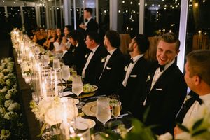 Groomsmen in black tuxedos laugh and smile at a candlelit wedding reception dinner table.