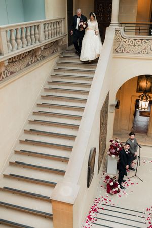 Bride descends a grand staircase with rose petal aisle at a luxurious wedding venue ceremony.