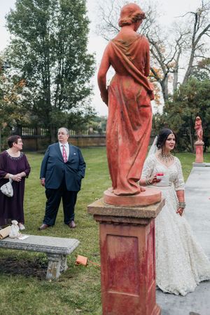 Indian bride in white embroidered lehenga stands near a red classical statue in a garden wedding setting.
