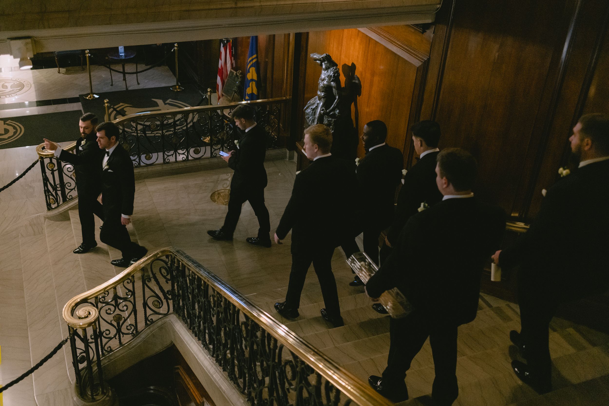 Groomsmen in black suits walk down an elegant staircase with ornate iron railings at a wedding venue.