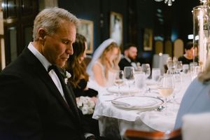 Father of the bride sits at a wedding reception table in a tuxedo, with the bride visible in the background.