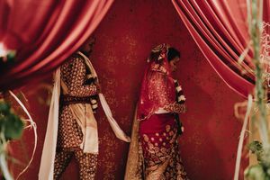 Bride and groom in ornate red and gold Indian wedding attire standing beneath red drapes.