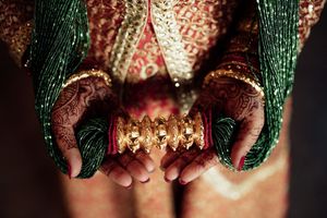 Indian bride displaying ornate henna-adorned hands with gold bangles and green beaded bridal jewelry.