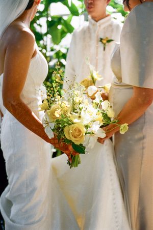 Bride holding elegant yellow rose and white flower bouquet during outdoor wedding ceremony.