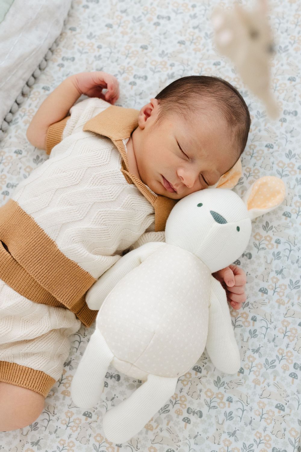 A sleeping baby rests peacefully on floral bedding while cuddling a white plush bunny toy.