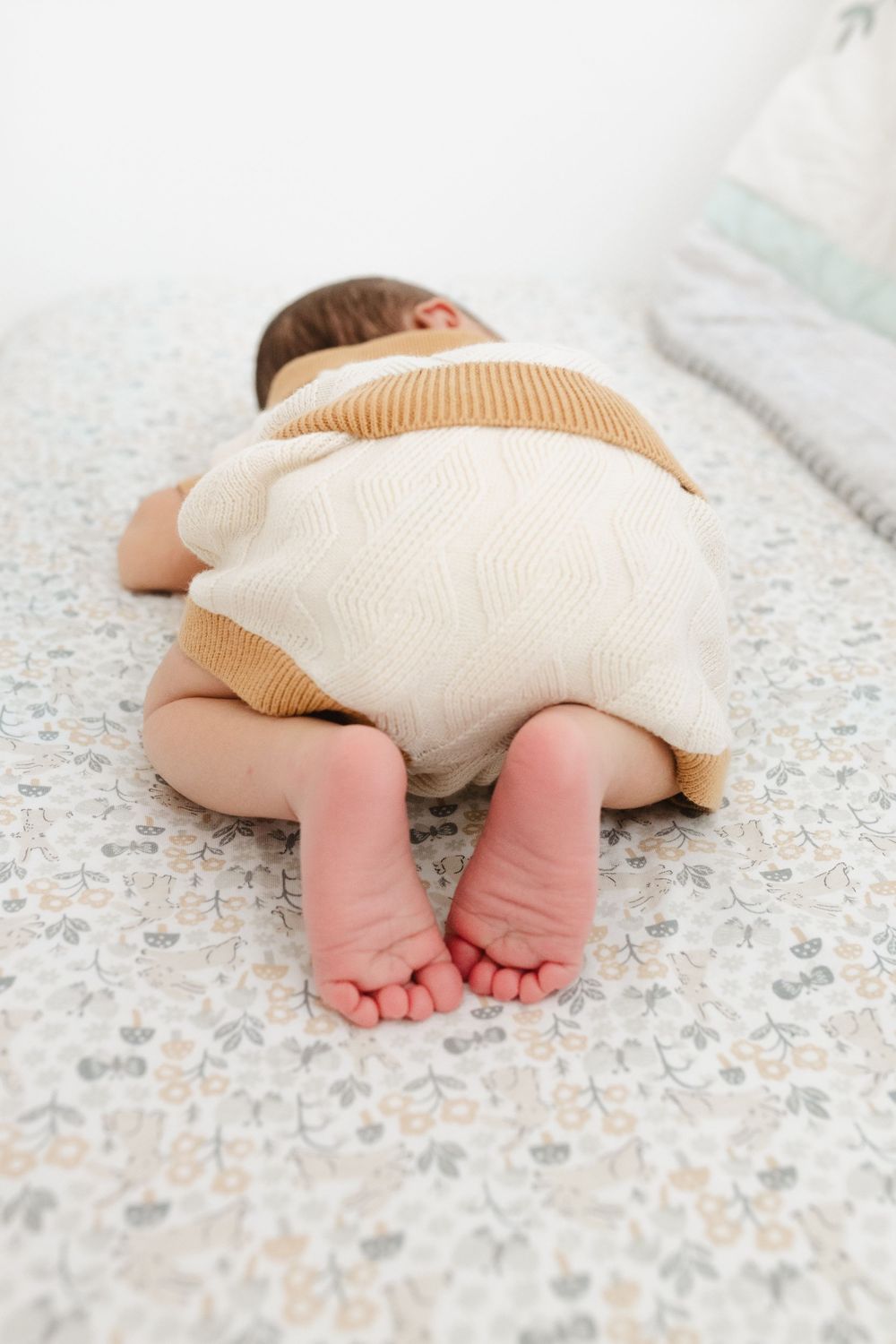 Baby wearing cream-colored outfit lying on patterned floor showing tiny feet.