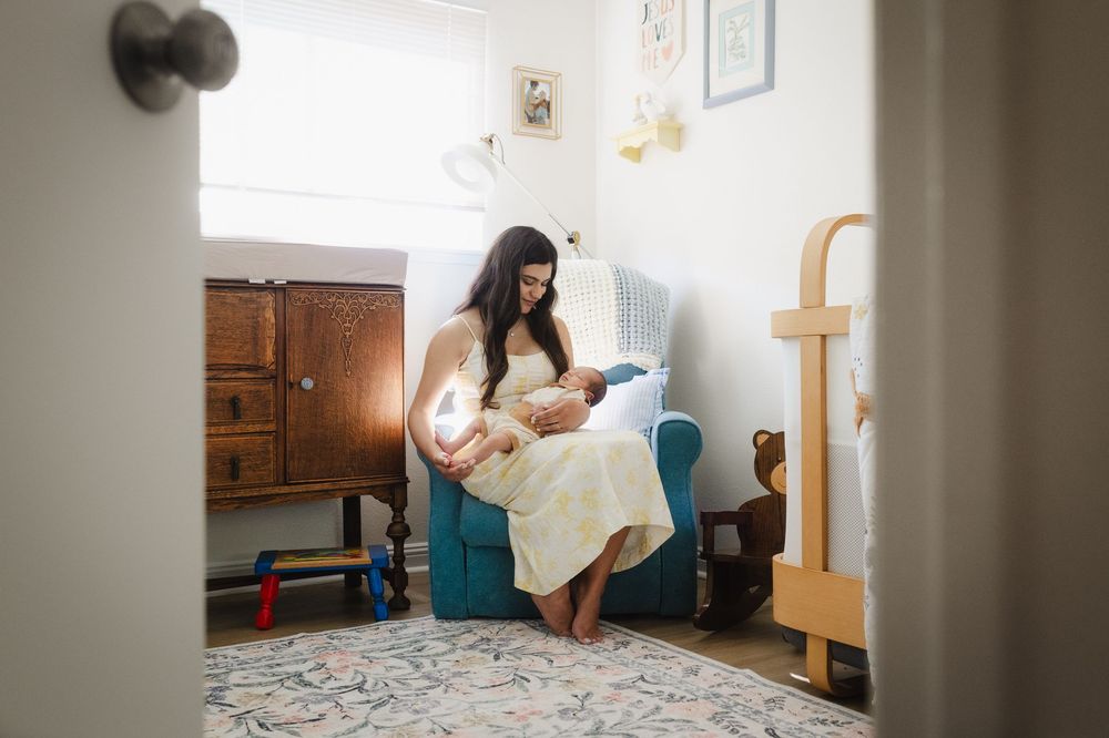 Parent sitting in blue chair in nursery corner reading to baby in white dress.