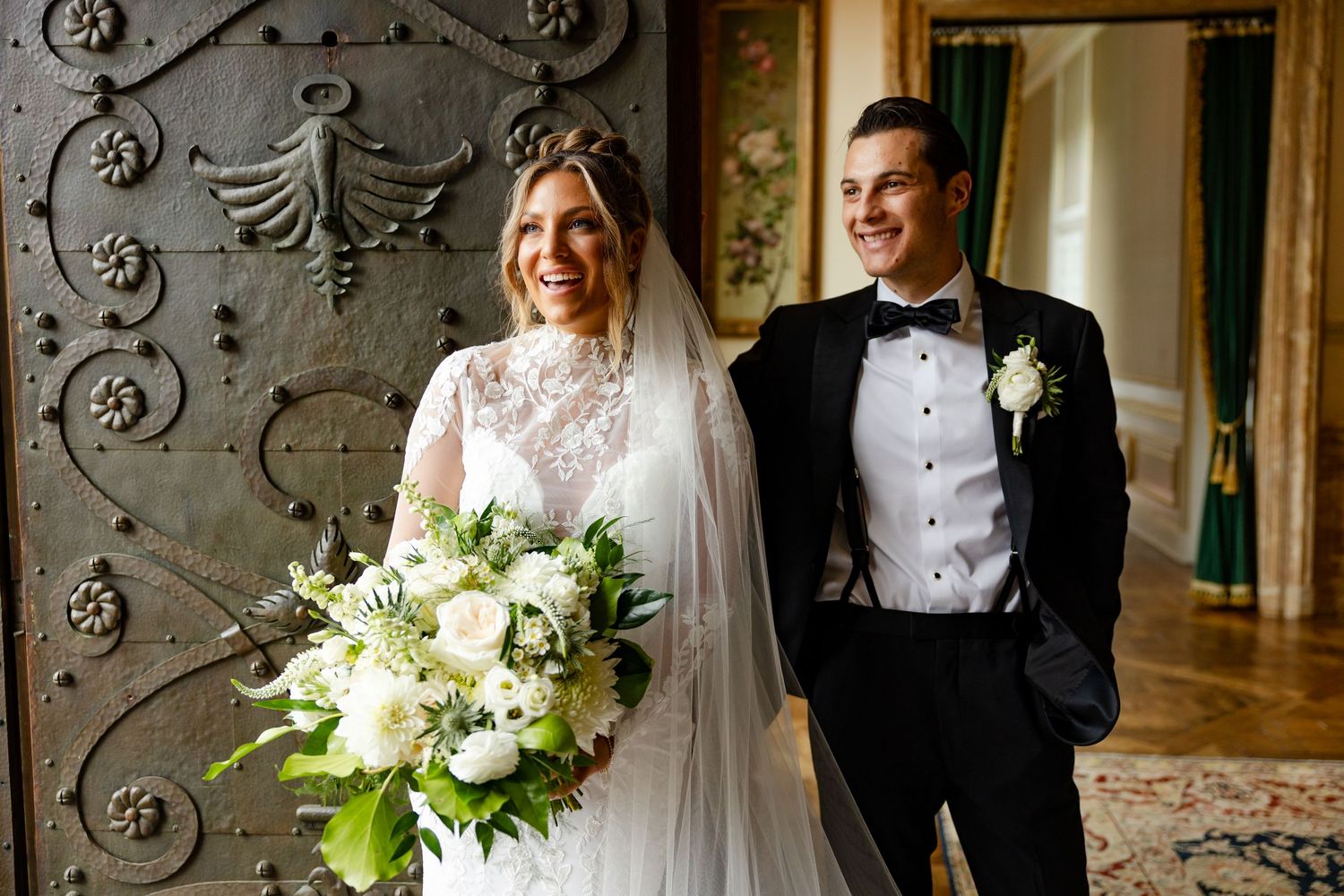 Elegant couple in formal wedding attire posing by ornate metal door with white floral bouquet.