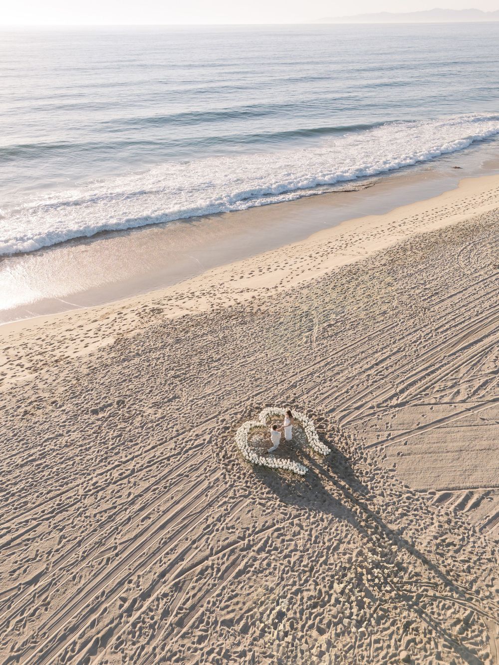Aerial view of beach waves rolling onto sandy shore with tire tracks creating patterns in the morning light.