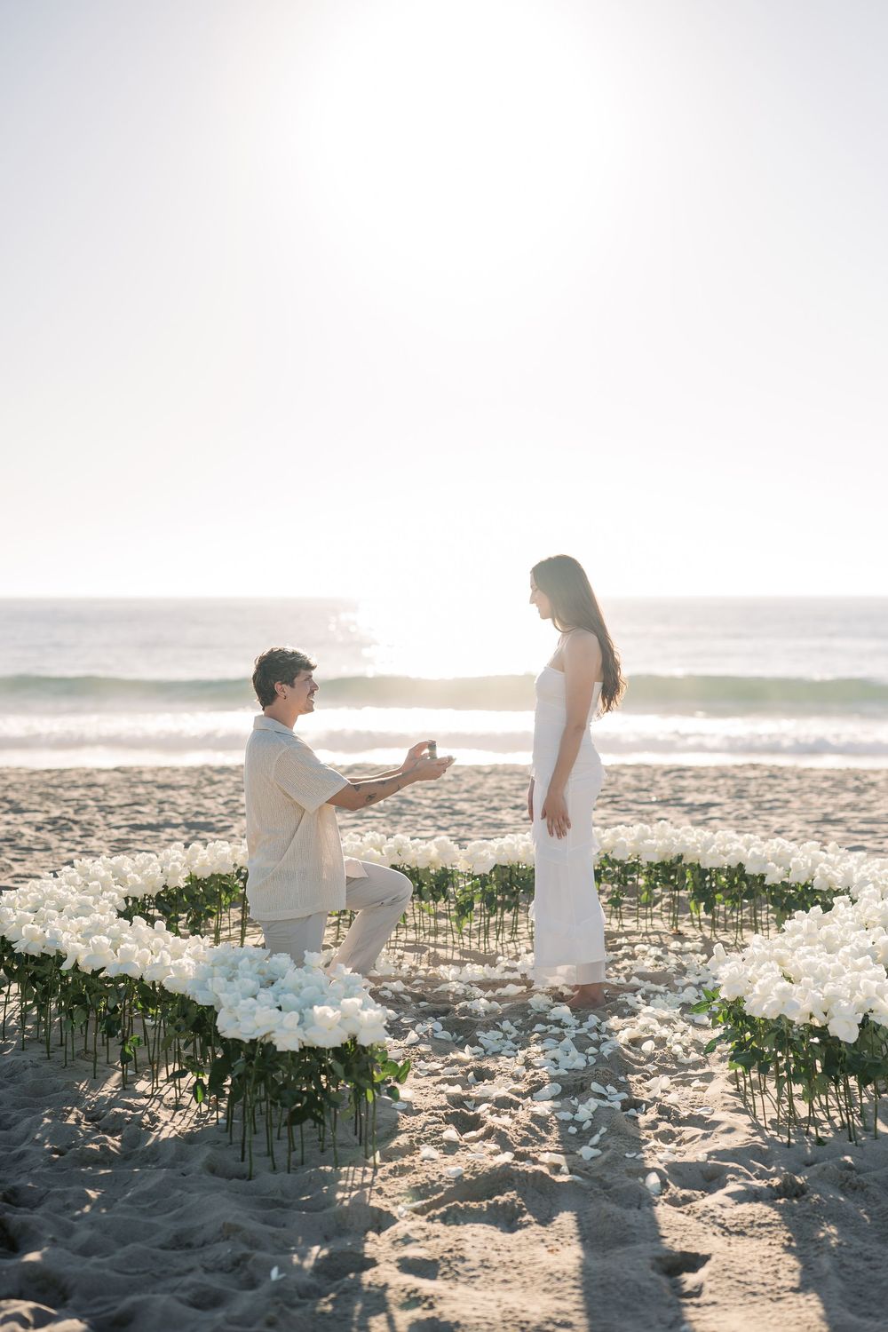 A romantic beach proposal at sunset with white flowers scattered on the sand creating a beautiful seaside moment.