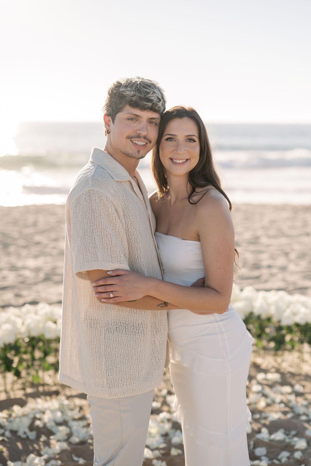 A couple in white outfits embraces on a sunny beach with ocean waves in the background.