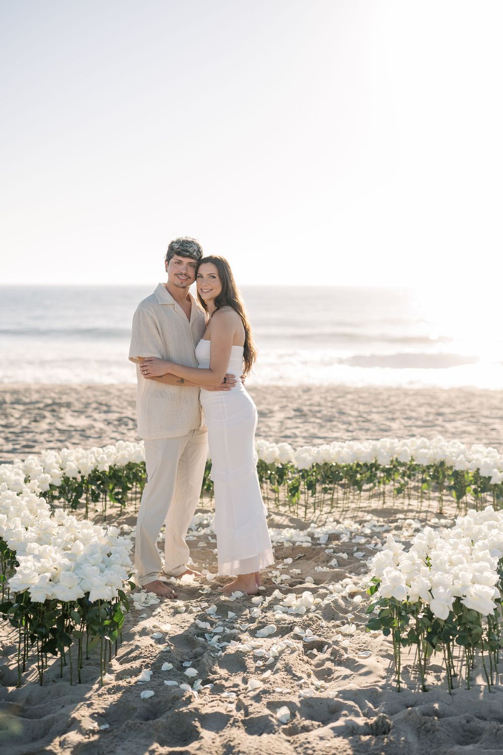 Romantic beach couple embraces at sunset surrounded by white flowers arranged in the sand during an intimate ceremony.