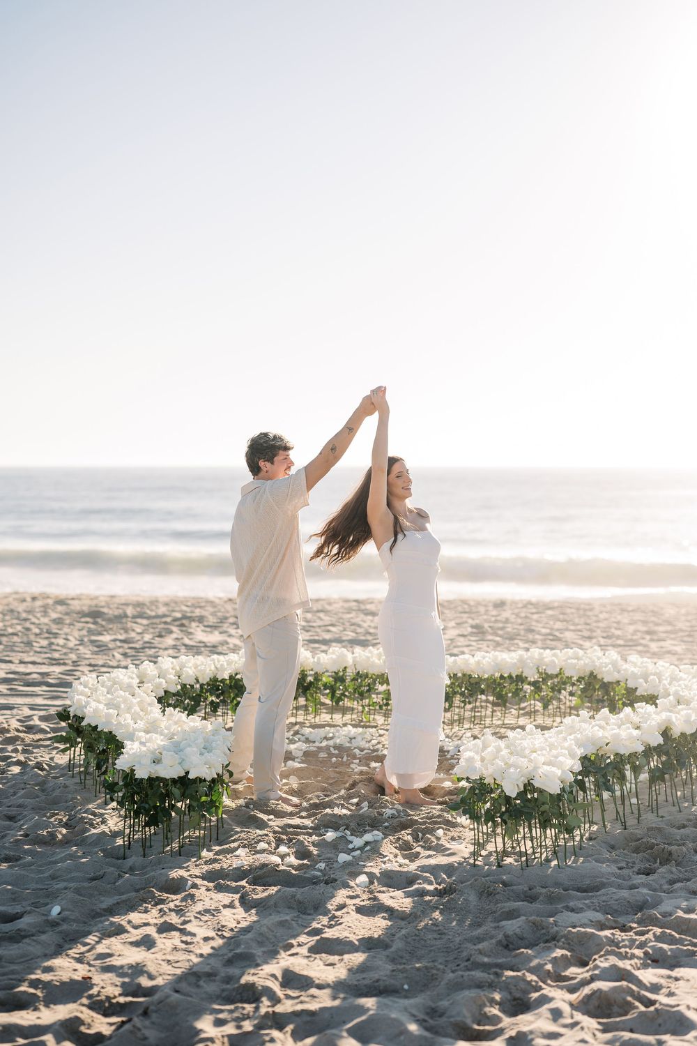 Dancing couple in white outfits celebrate on beach with floral decorations.