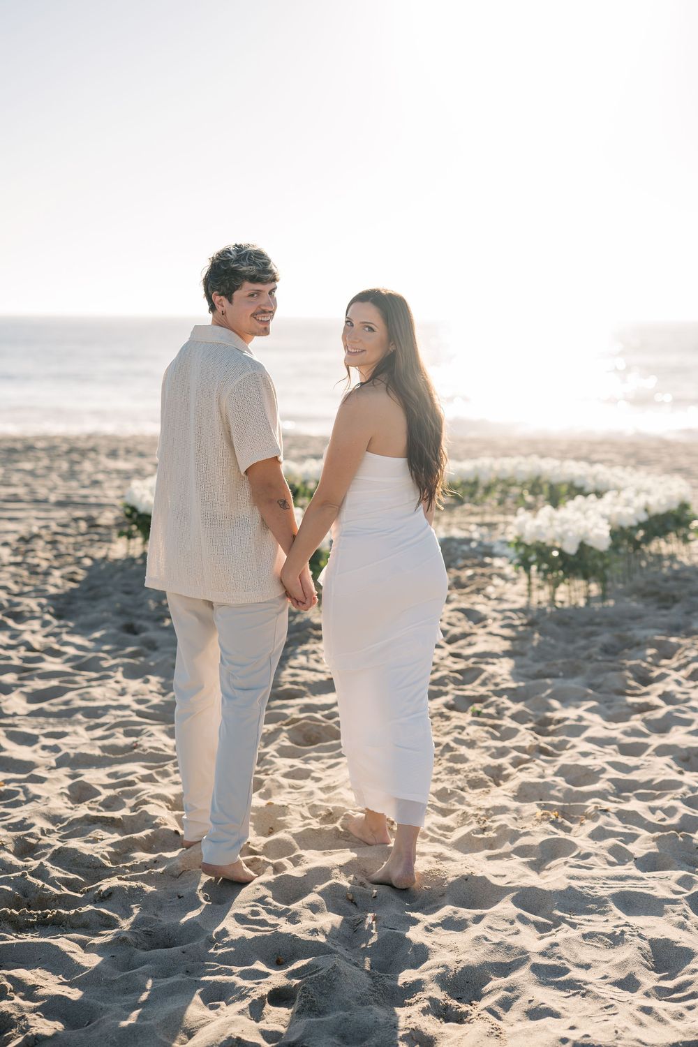 Couple in matching white outfits hold hands on sandy beach with flowers.