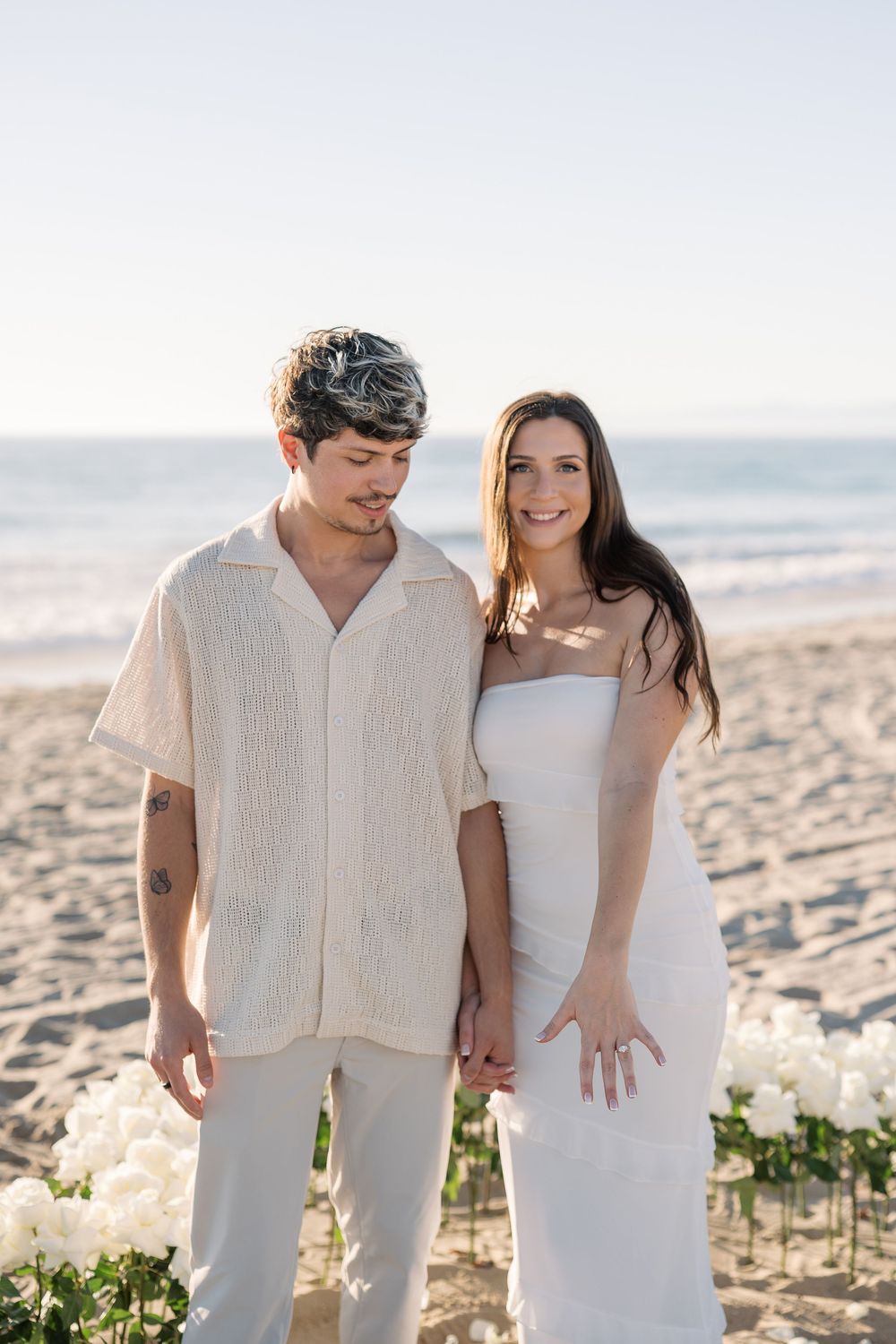 A romantic beach photoshoot featuring a couple in matching light-colored attire against a sandy backdrop.