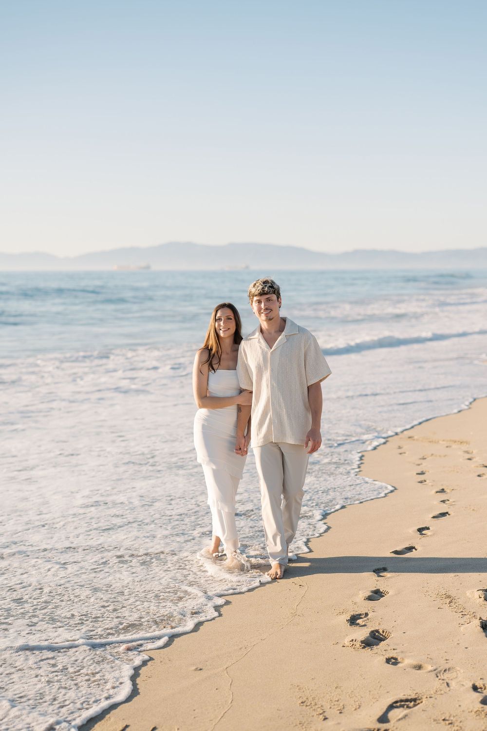 Couple strolls along beach at sunset leaving footprints in sand.