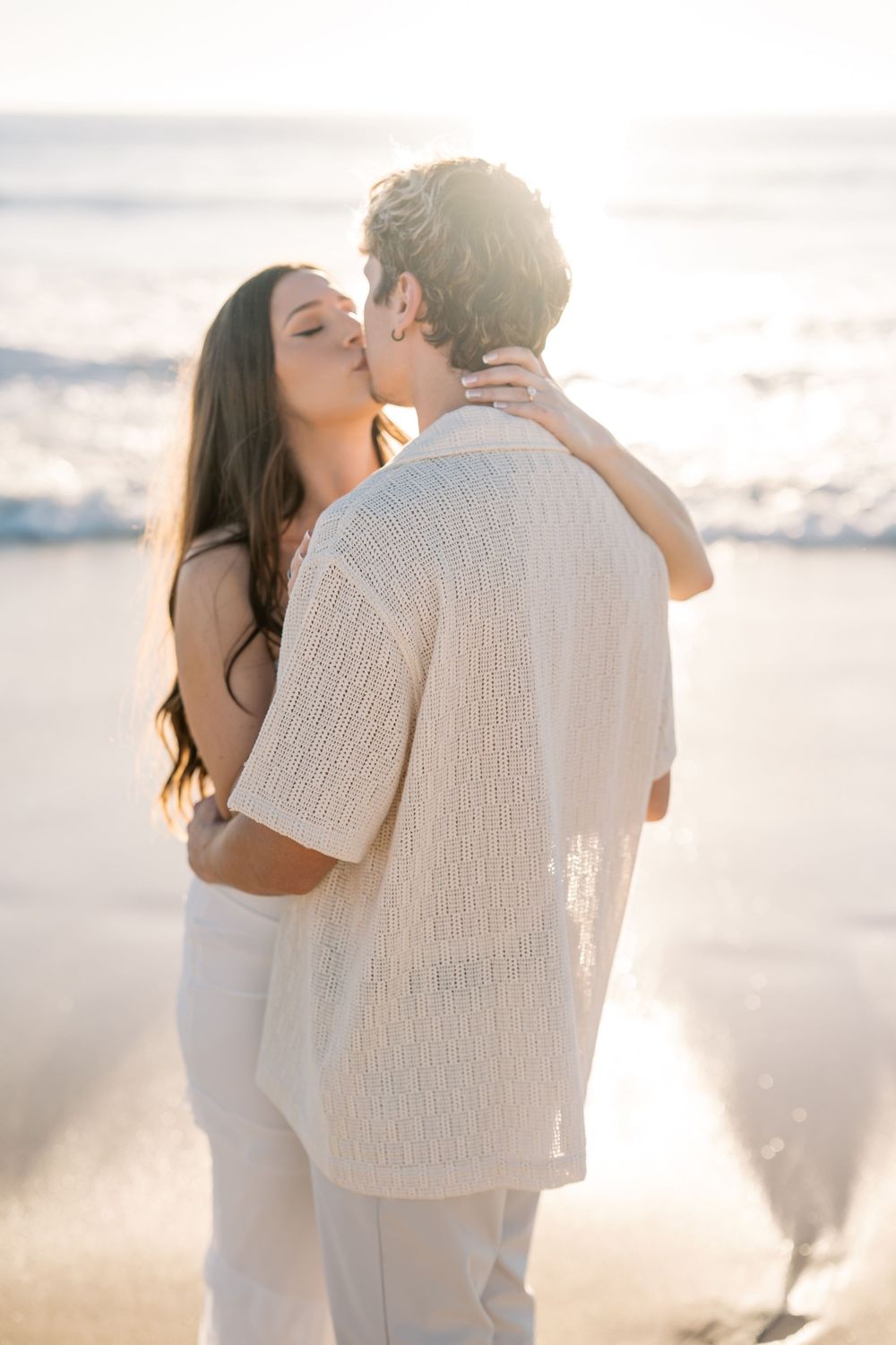 A romantic couple in white clothing shares intimate moments on a sunlit beach with waves in the background.