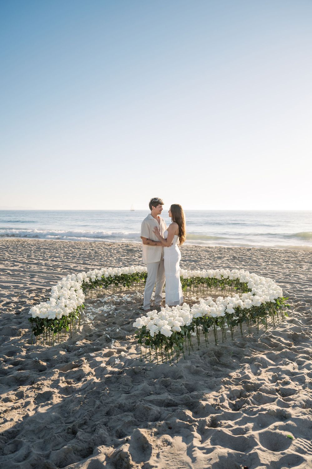 A couple stands inside a heart shape made of white flowers on the beach at sunset.