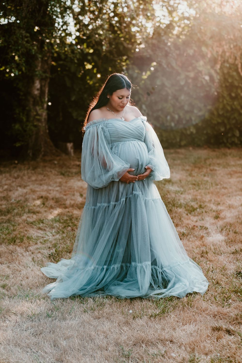 Expectant mother in light blue off-shoulder tulle dress cradling baby bump at sunset.