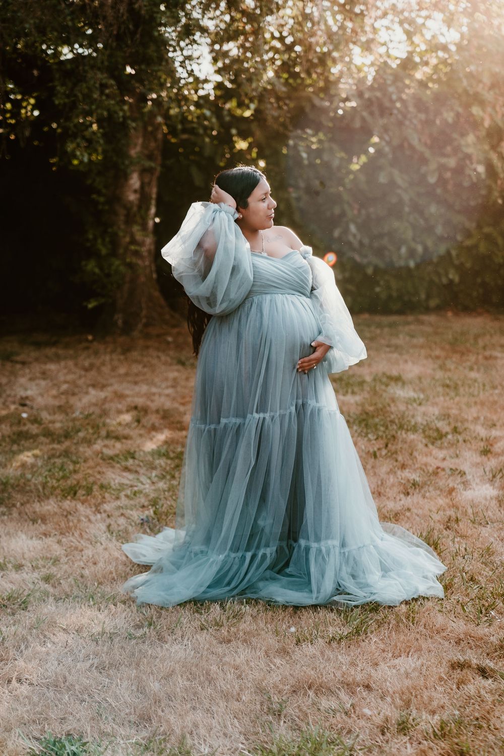 Pregnant woman in dusty blue tulle maternity dress posing in sunlit outdoor field.