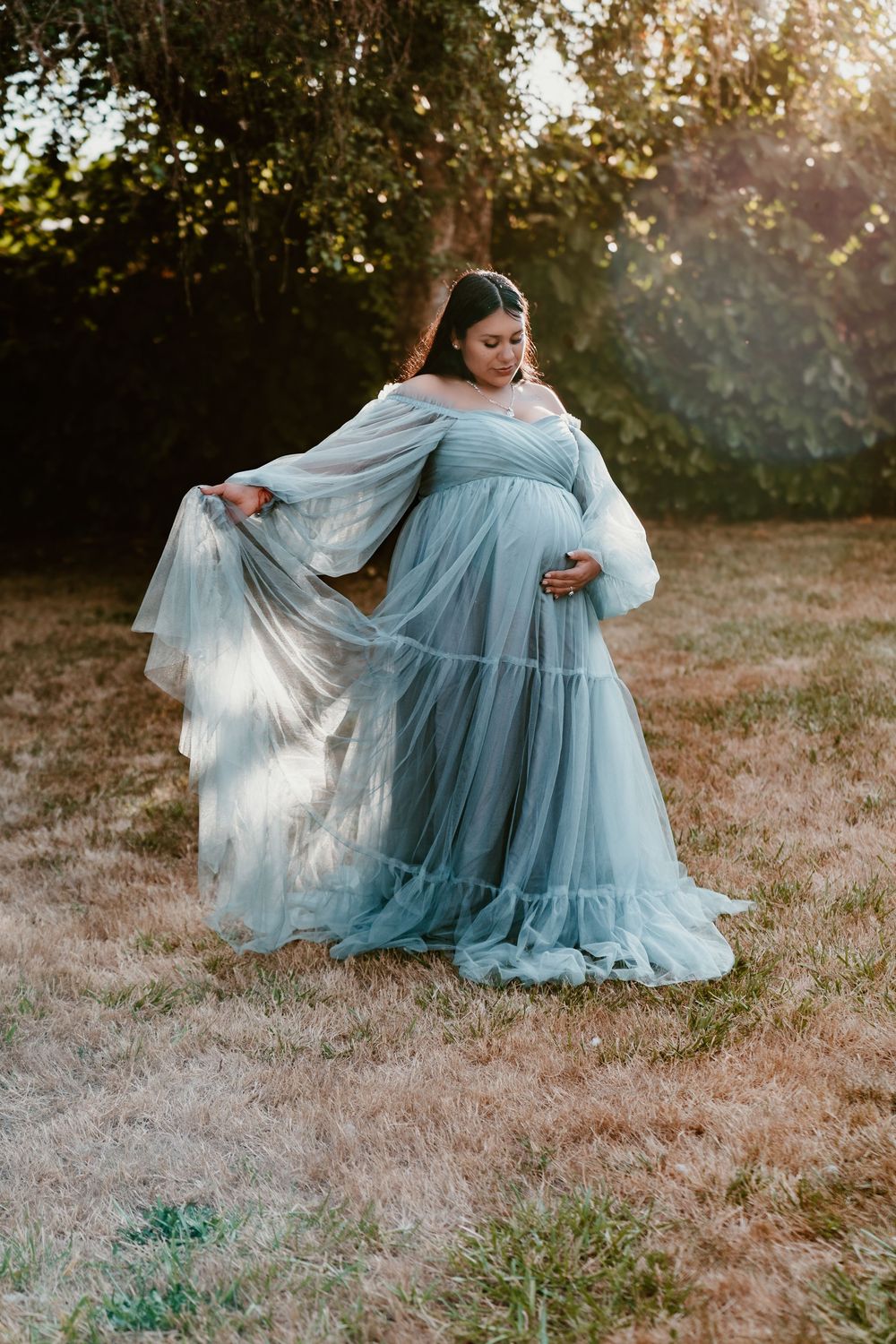 Pregnant woman in flowing teal tulle maternity gown posing outdoors at golden hour.