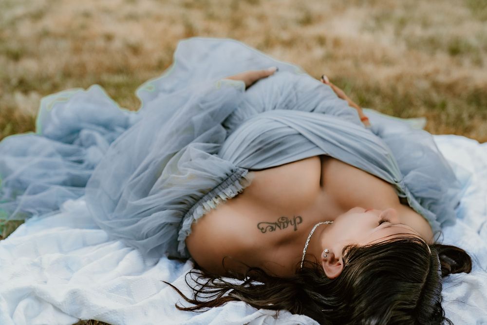 Woman in flowing blue tulle dress lying on white blanket with script tattoo visible.