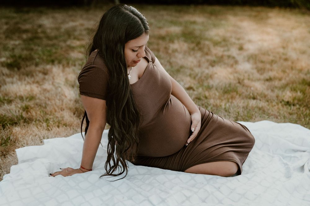 Pregnant woman in brown dress sitting on white blanket cradling baby bump outdoors.