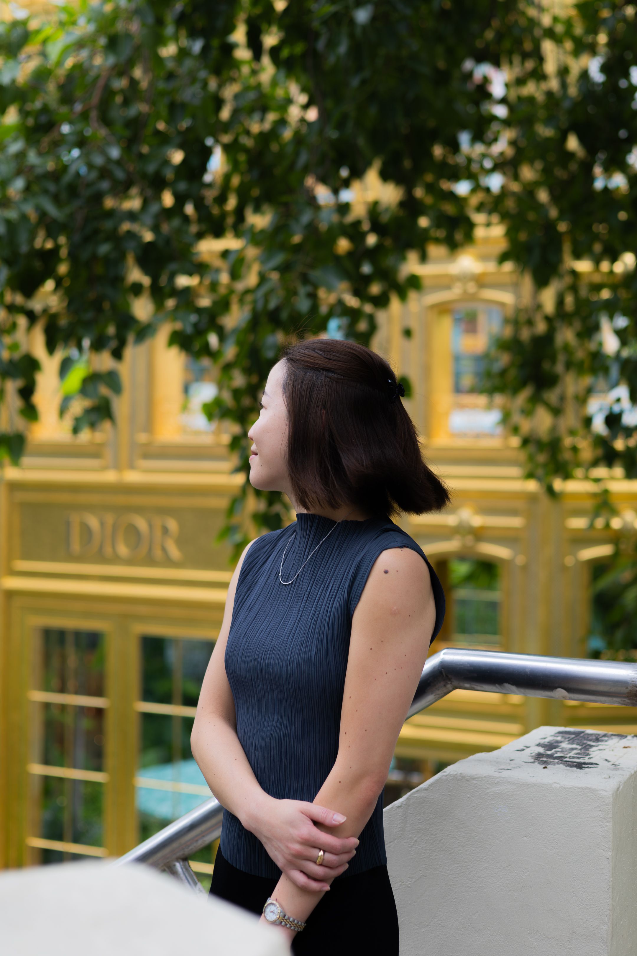 Person in sleeveless black top standing outside a Dior store with yellow facade and green foliage overhead.