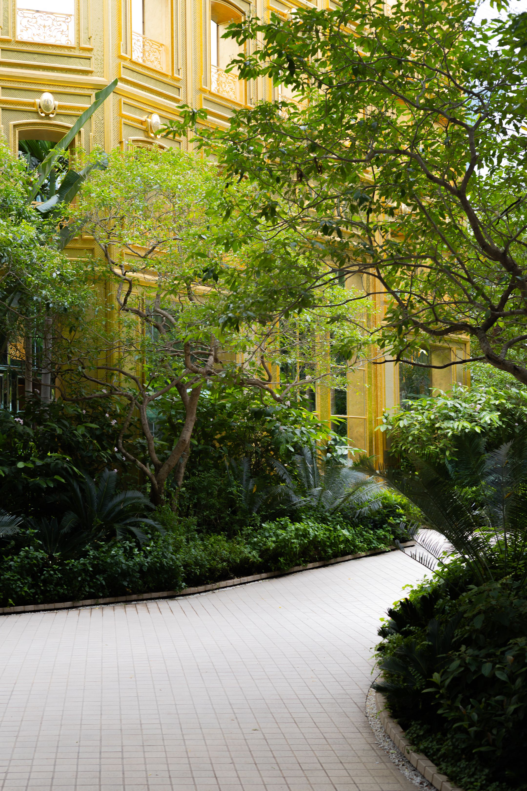 A curved garden path with white tiles leads past green shrubs and trees alongside a yellow building exterior.