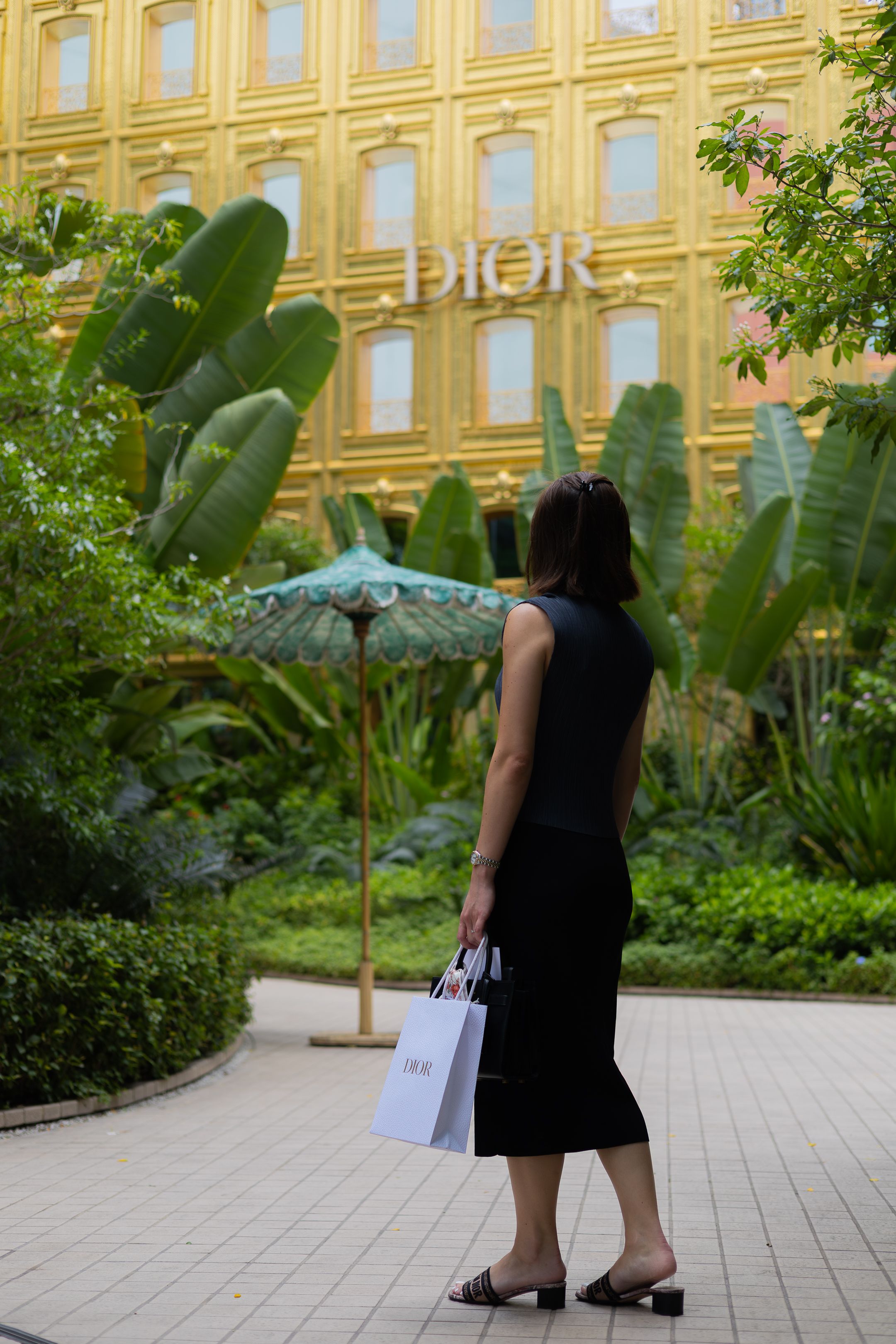 A person in a black dress carries shopping bags while standing in a lush garden courtyard with a yellow building facade.