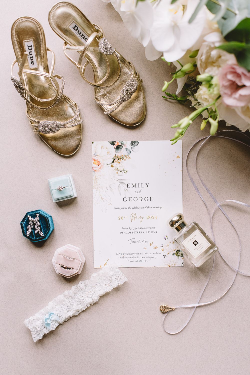 Elegant wedding details flatlay with gold sandals, white lace garter, perfume bottles, and floral wedding invitations.
