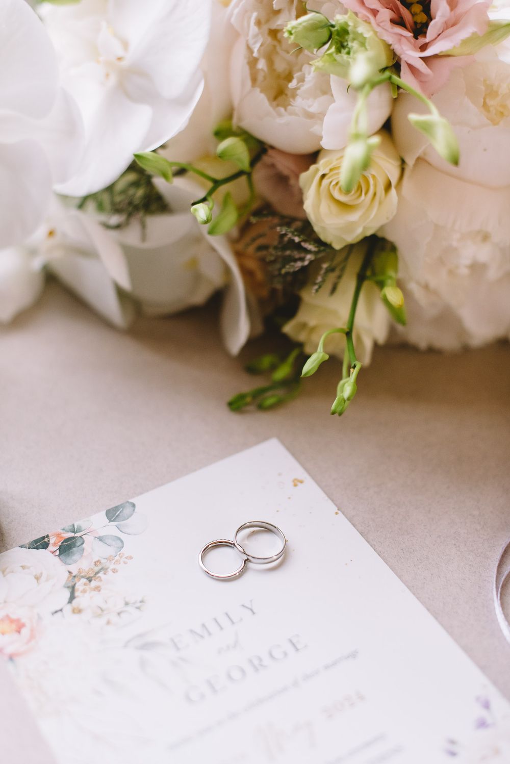 Wedding rings rest on delicate floral stationery surrounded by soft pink and white roses and greenery.
