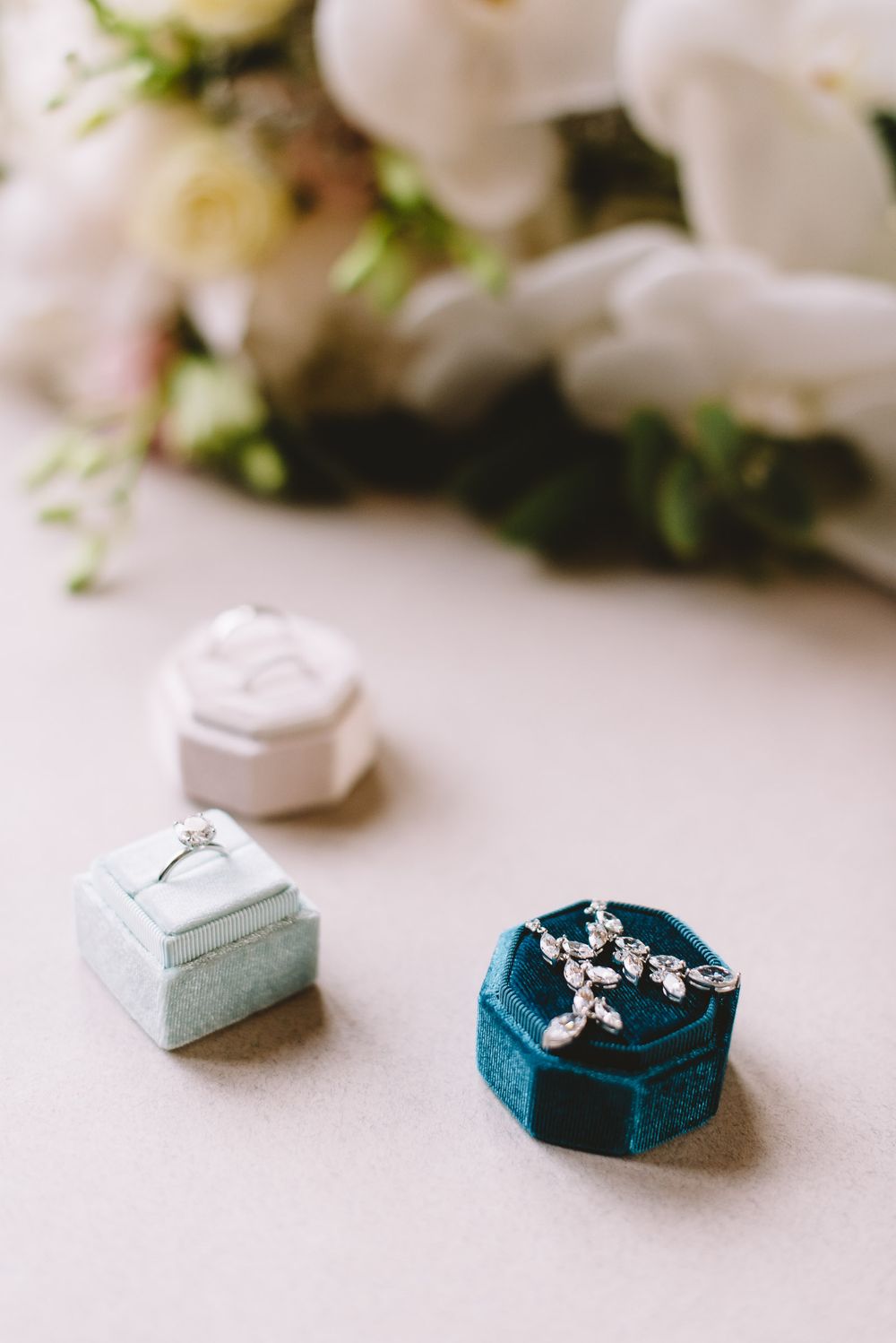 Three velvet ring boxes in teal and white colors with delicate jewelry displayed against a light background.
