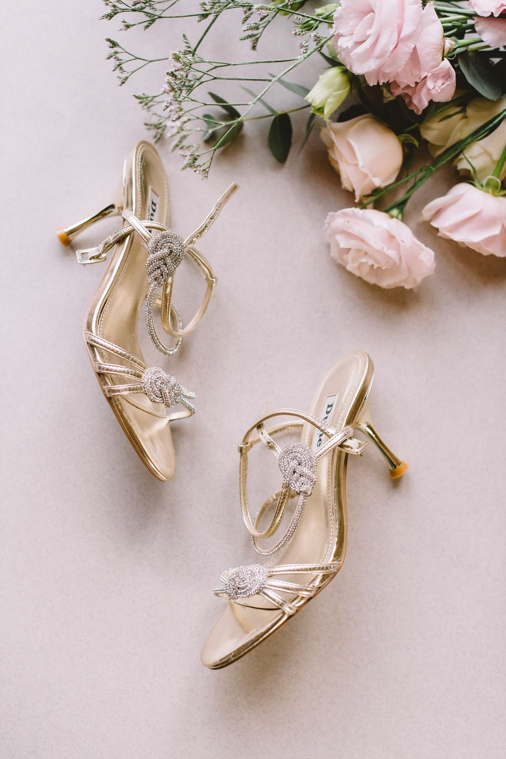 Elegant silver bridal heels with crystal embellishments arranged with pink roses on a light neutral background.
