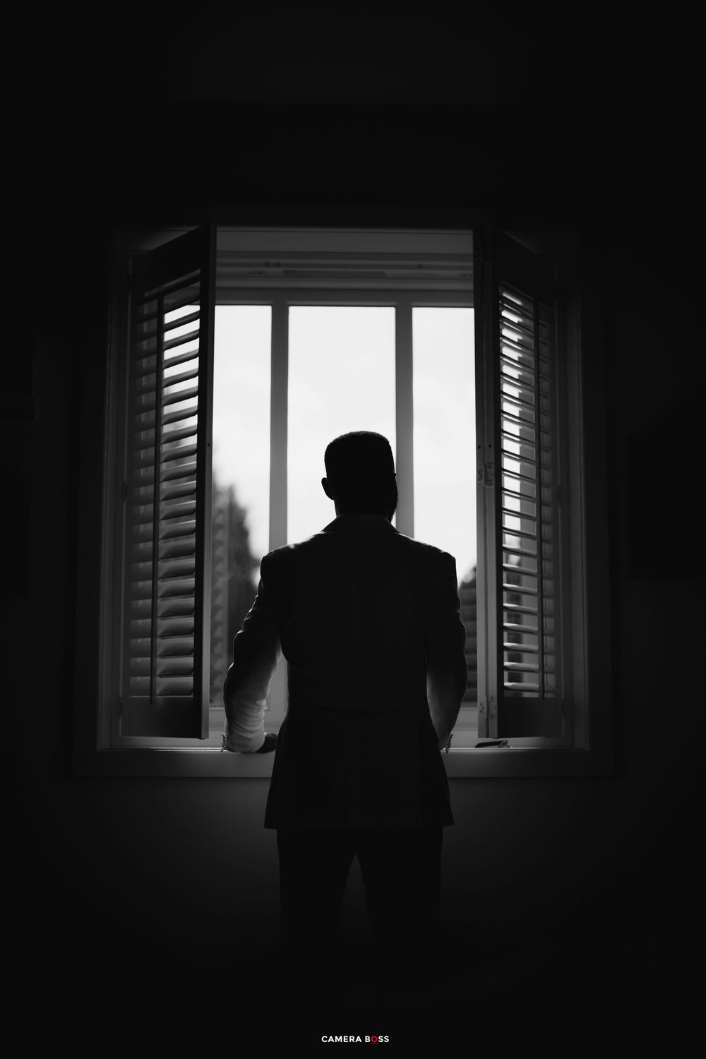 A dark silhouette stands by a window with slatted shutters in a moody black and white composition.