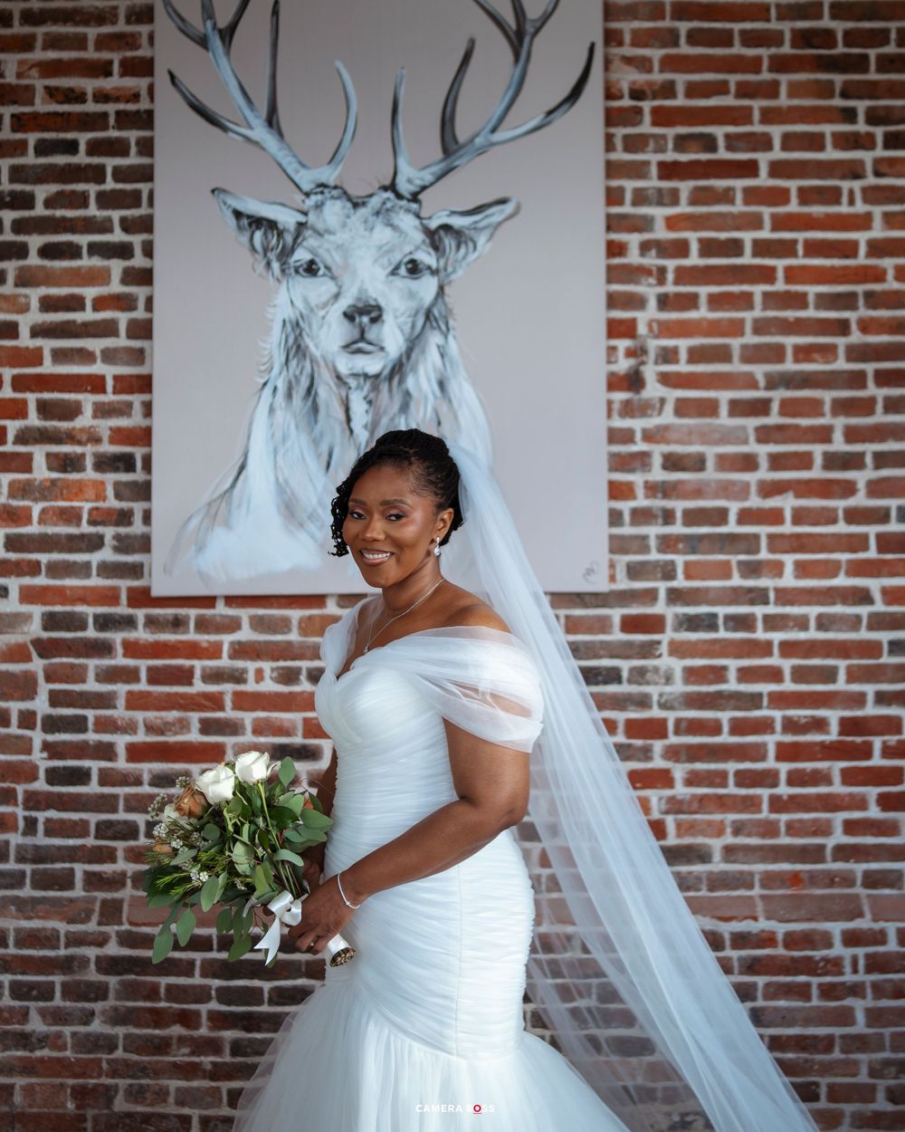 A bride in a white strapless wedding gown poses against a brick wall with deer artwork.