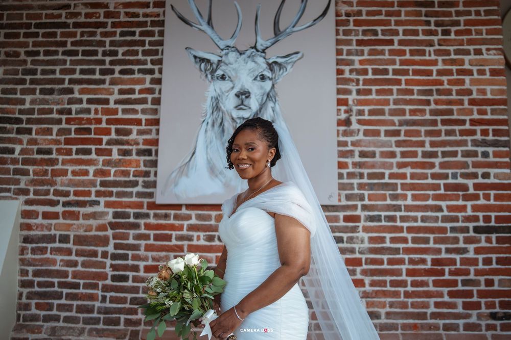A bride in a white dress poses in front of a brick wall with a large deer artwork behind her.